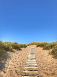 View of footpath leading towards sand against clear blue sky