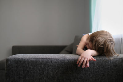 European toddler child sits and gets bored on gray sofa in bright living room, minimalist interior