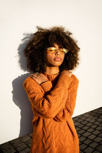 Full length of thoughtful cute ethnic woman with curly afro hair wearing vivid orange sweater with closed eyes against white modern concrete wall background