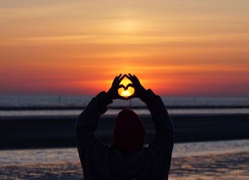 Woman making heart shape at beach against sky during sunset