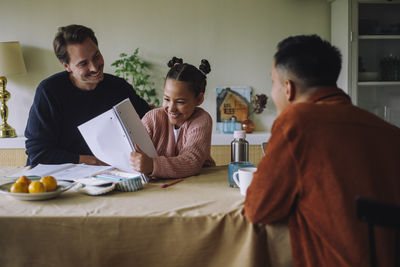 Happy daughter showing notebook to gay father at dining table in home