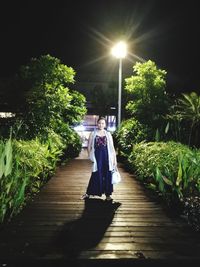 Portrait of woman standing on illuminated walkway amidst trees against sky