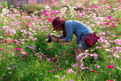 Side view of woman photographing with camera while standing amidst flowering plants