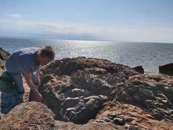 Rear view of boy on rock at beach against sky