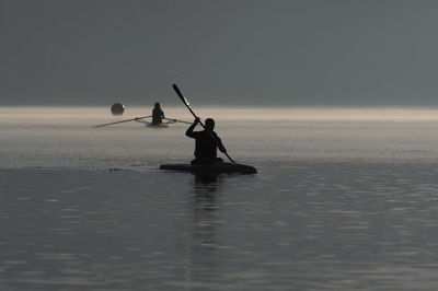 Silhouette people oaring boats on sea against sky