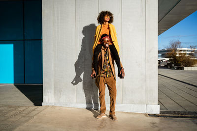From below of modern african american guy in sunglasses and jacket carrying trendy black girlfriend on shoulders standing on street looking away