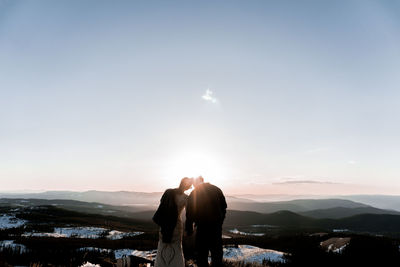 Rear view of woman and man standing on landscape against sky during sunset on mountaintop