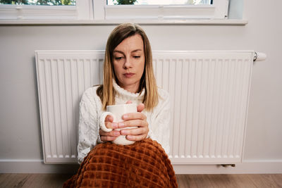 Worried woman sit near heating radiator under blanket with cup of tea