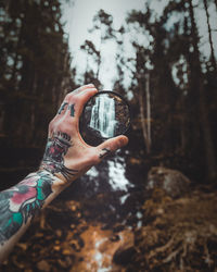 Cropped tattooed hand holding glass in front of waterfall