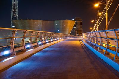 Illuminated footbridge by buildings against sky at night