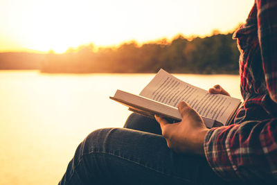 Woman reading book while sitting by lake against sky during sunset
