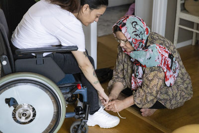 Mother tying shoelace of daughter sitting on wheelchair at home