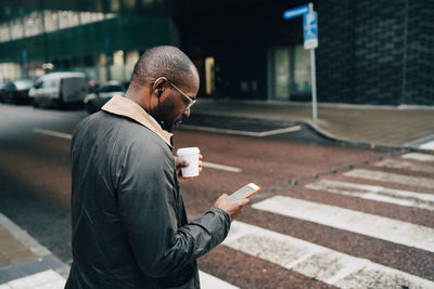 Side view of businessman with coffee using mobile phone while crossing road in city