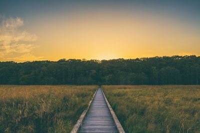 Boardwalk amidst field against sky during sunset