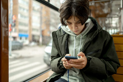 Boy looking at mobile phone sitting by window in tram