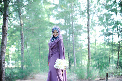 Bride in traditional clothing holding flower bouquet while standing against trees in forest