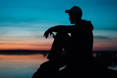 Silhouette man sitting on rock at beach against sky during sunset