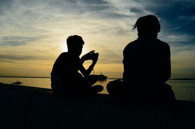 Silhouette men sitting on beach against sky during sunset