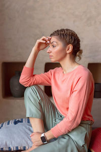 Pensive beautiful woman in coral sweatshirt and gray pants is sitting in the room