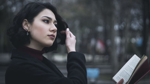 Young woman looking away holding book while sitting in city