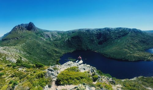 High angle view of man sitting on cliff over lake by mountain against clear sky