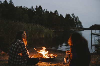 Rear view of female friends talking by fire pit against lake during sunset