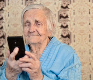 Happy elderly 90-year-old woman with glasses wearing a blue jacket smiles using a smartphone.