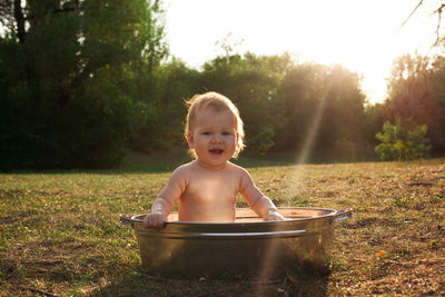 Cute little kid sits in a basin of water in nature and has rays of the setting sun, golden bathe. 