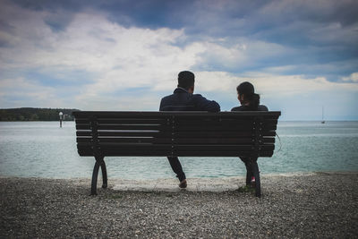 Rear view of men sitting on bench by sea against sky