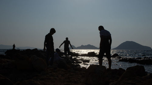 Silhouette people on rocks by sea against clear sky