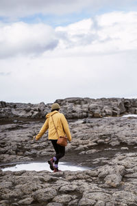 Back view of anonymous young female tourist in yellow raincoat and hat walking on rough volcanic terrain near small ponds under cloudy sky in iceland