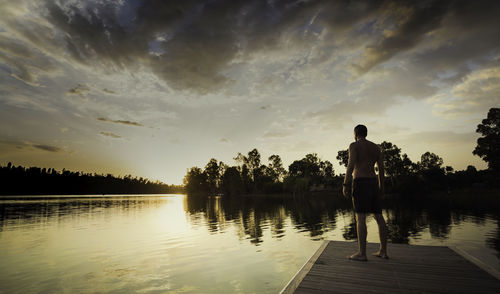 Young man standing without shirt looking at the sunset from the wooden jetty