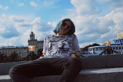 Portrait of teenage boy wearing sunglasses while sitting on railing in city against cloudy sky