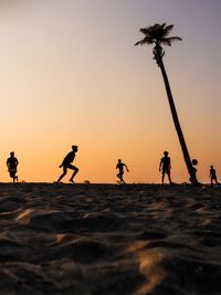 Silhouette male friends playing soccer at beach against clear sky during sunset
