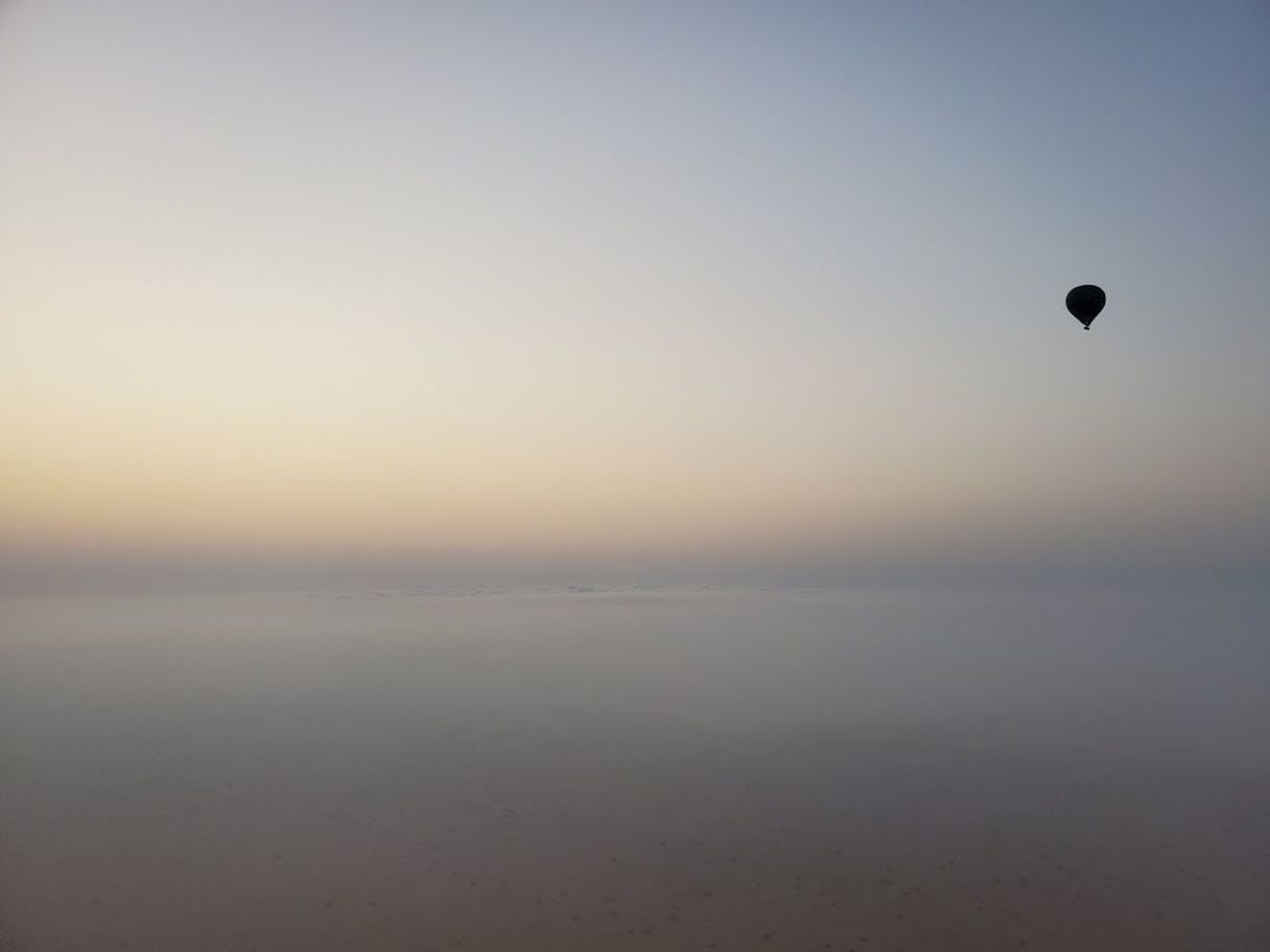 sky, hot air balloon, mid-air, flying, horizon, balloon, tranquility, scenics - nature, nature, beauty in nature, sunrise, tranquil scene, air vehicle, aircraft, dawn, morning, environment, water, adventure, sea, transportation, vehicle, fog, copy space, no people, outdoors, travel, land, landscape, idyllic, cloud, hot air ballooning, sun, reflection, sunlight, horizon over water, blue, day, distant