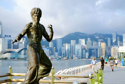 People walking by bruce lee statue at tsim sha tsui against cloudy sky