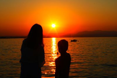 Silhouette boy and girl standing by sea against orange sky during sunset