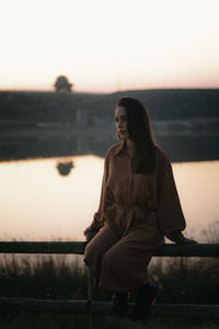 Young woman looking away while sitting on railing against sky during sunset