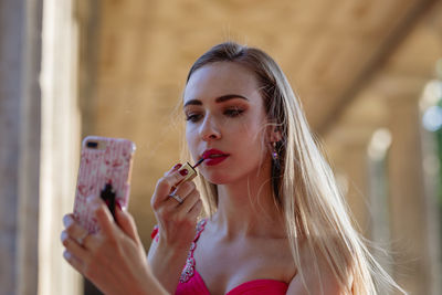 Young woman in pink evening gown applying lipstick while looking at smart phone outdoors