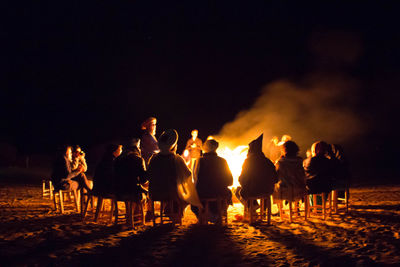 People sitting side by side at campfire against clear sky during night