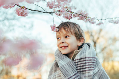 Portrait of a funny little boy wrapped in a blanket enjoying cherry blossoms in a city park.