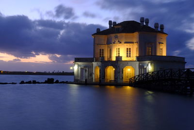 Illuminated building by sea against sky at sunset