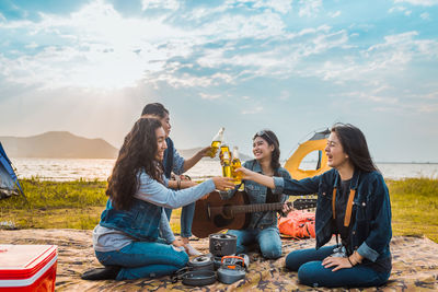 Friends toasting beer bottles while camping at lakeshore against sky