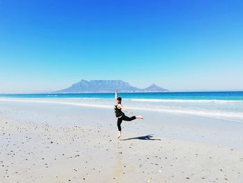 Cheerful woman jumping on beach against clear blue sky