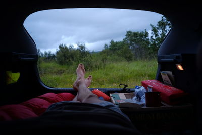 Low section of man sleeping in car at campsite against cloudy sky