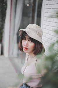 Portrait of young woman wearing hat while sitting against wall