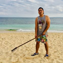 Portrait of man holding rake standing at shore of beach against sky