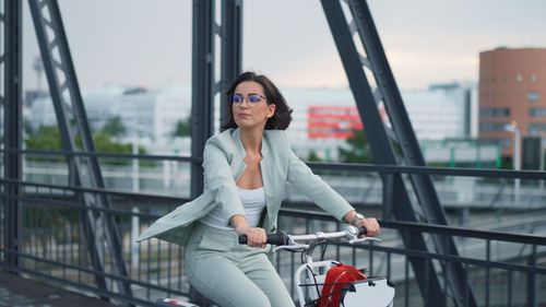 Young happy businesswoman enjoys riding bike on citybridge with beautiful cityscape in the morning