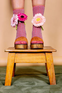 Crop anonymous bride in white dress and high heeled shoes with colorful flowers standing on wooden stool against beige background in studio