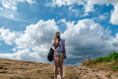 Full length of woman photographing while standing on field against cloudy sky